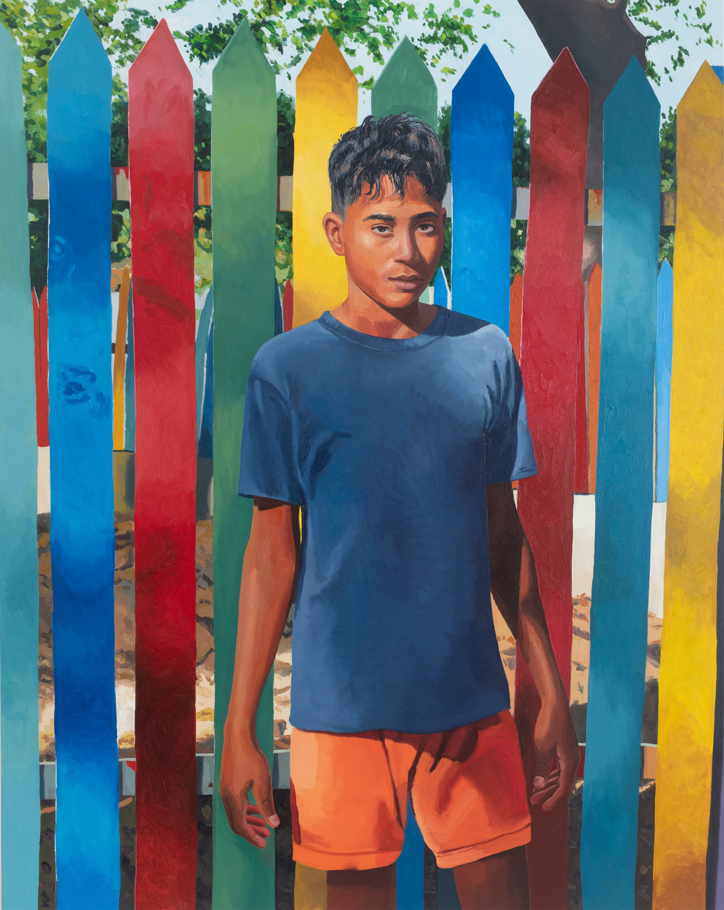 A young, brown-skinned masculine stands before a colorful fence. 